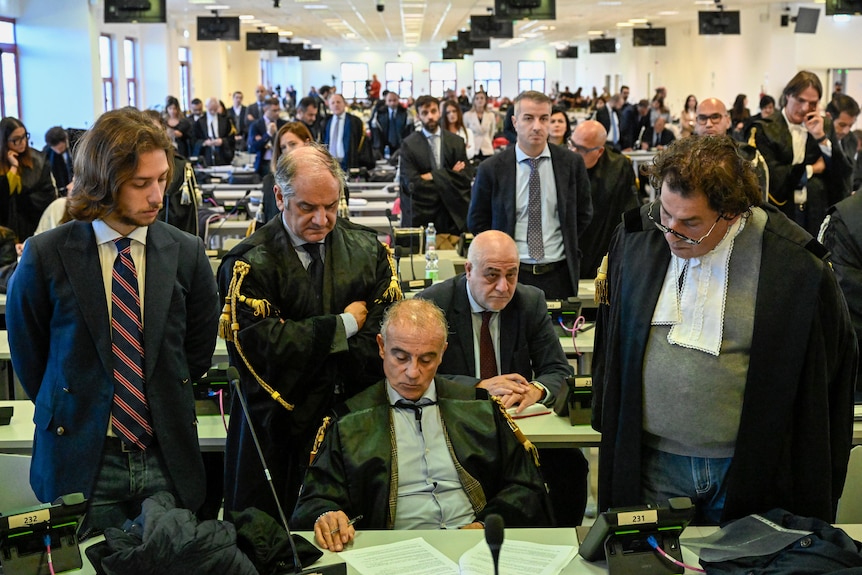 Men in suits look concerned as the stand and sit around a table