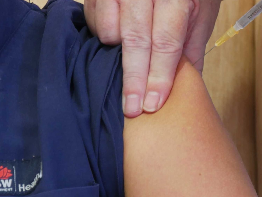 A syringe is inserted into the left shoulder of a uniformed female nurse, who is seated and smiling.