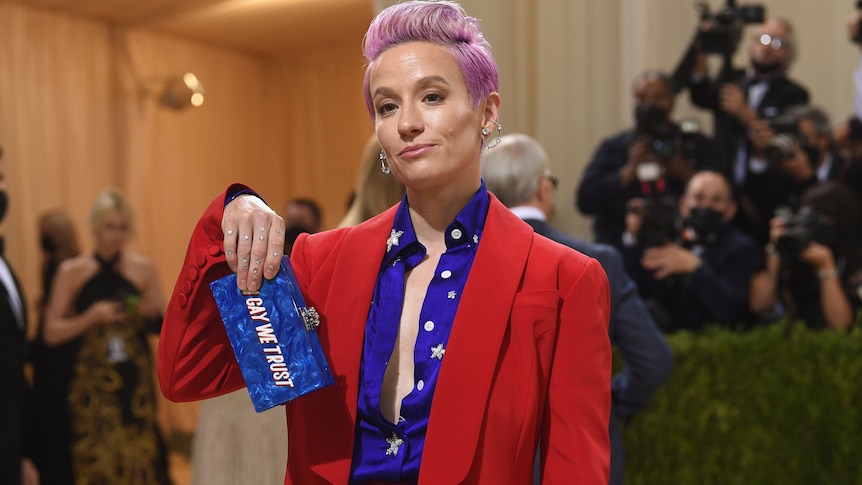 Megan Rapinoe holds up a clutch that reads "in gay we trust".