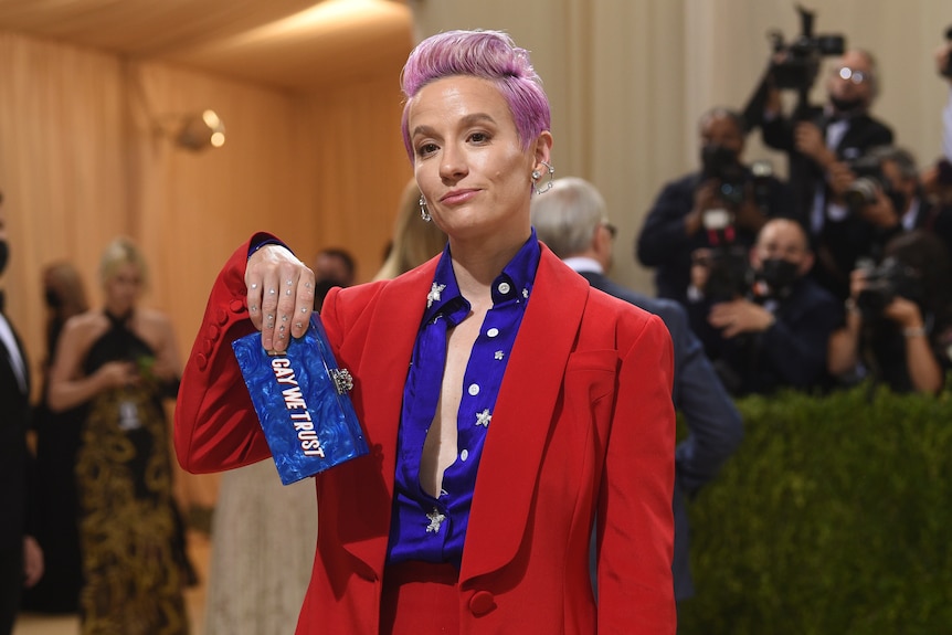 Megan Rapinoe holds up a clutch that reads "in gay we trust".