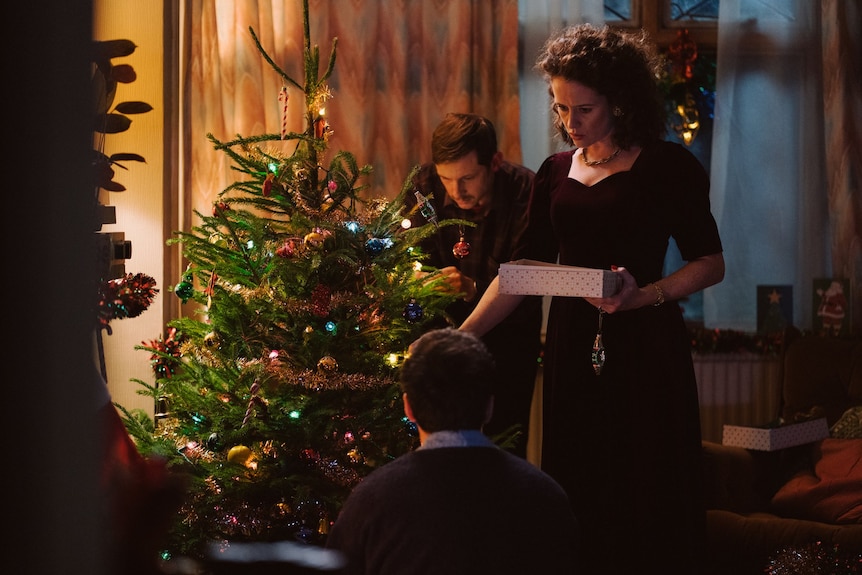 Claire Foy and Jamie Bell decorate a Christmas tree in an 80s living room, with Andrew Scott on the floor facing them.