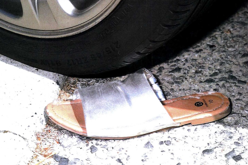 A close-up shot of a woman's silver slip-on shoe lying next to the wheel of a car near a kerb on the road.