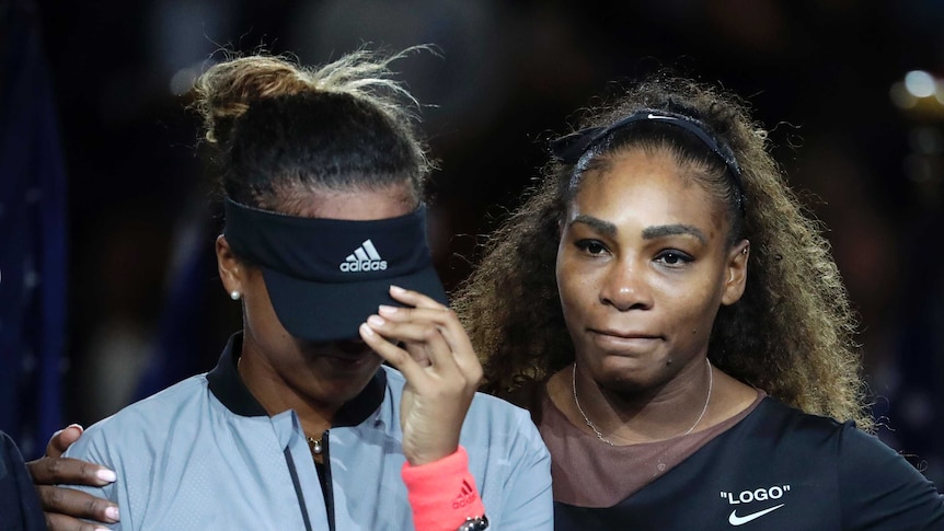 Naomi Osaka pulls her visor down as she's hugged by Serena Williams