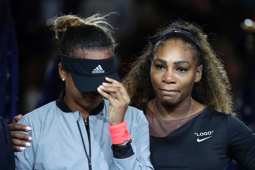 Naomi Osaka pulls her visor down as she's hugged by Serena Williams