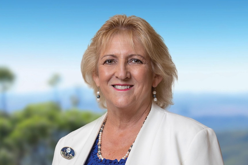 A woman with short blonde hair smiles. She is wearing a white blazer