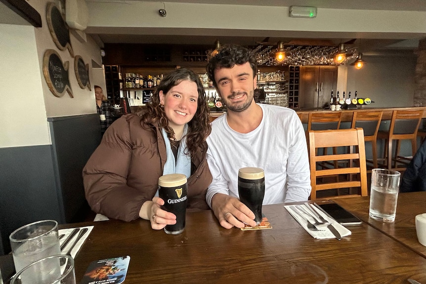 A young woman and young man sit smiling in a pub, each with a pint of Guinness.