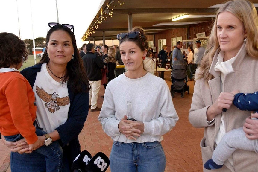 Three women stand in front of a crowd of people, with one holding a toddler and one holding a baby.