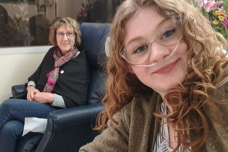 A young woman smiles at the camera as her mother looks on from an armchair behind her