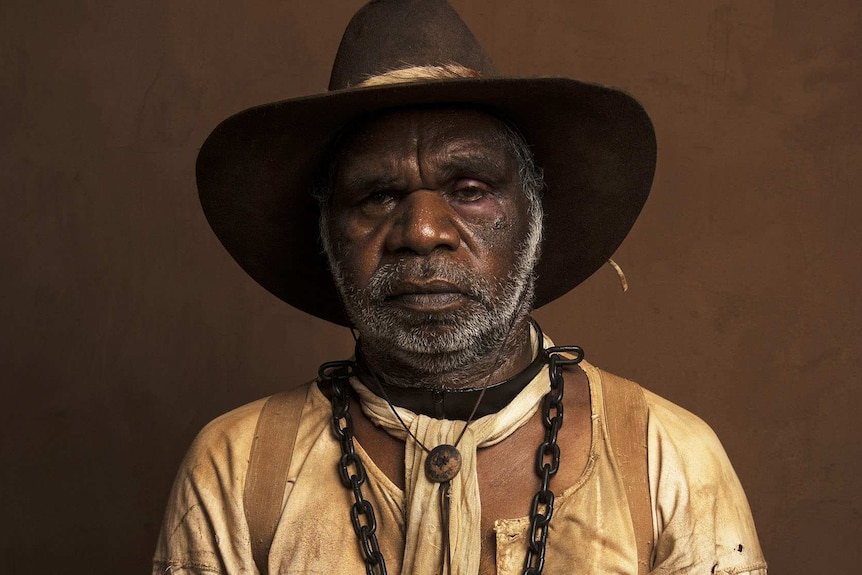 The actor, in tattered period garb and with iron chains around his neck stares into the camera forlornly against a brown backing