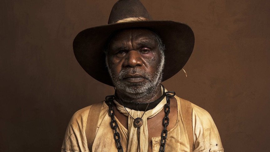 The actor, in tattered period garb and with iron chains around his neck stares into the camera forlornly against a brown backing