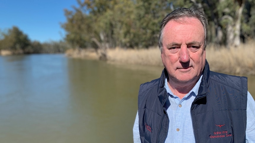 A man wearing a vest over a blue shirt stands before a section of river with the bush in the background