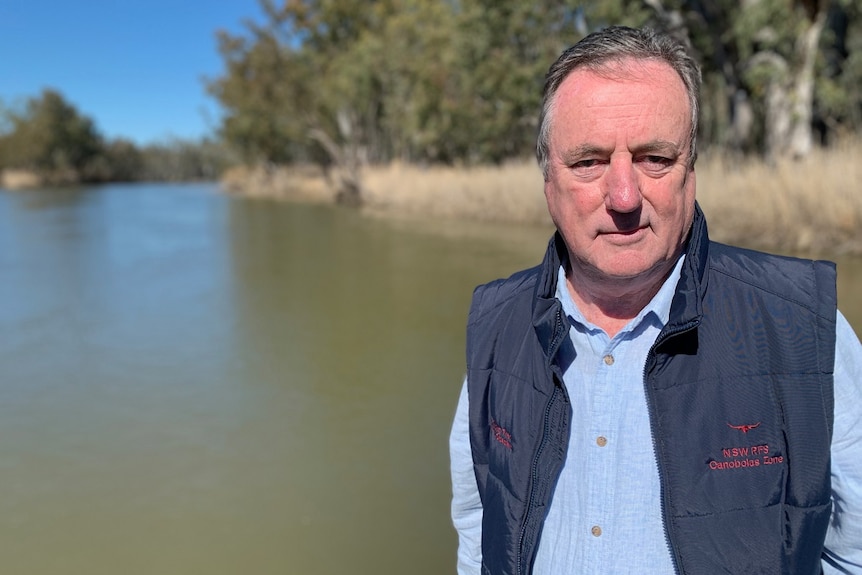 A man wearing a vest over a blue shirt stands before a section of river with the bush in the background