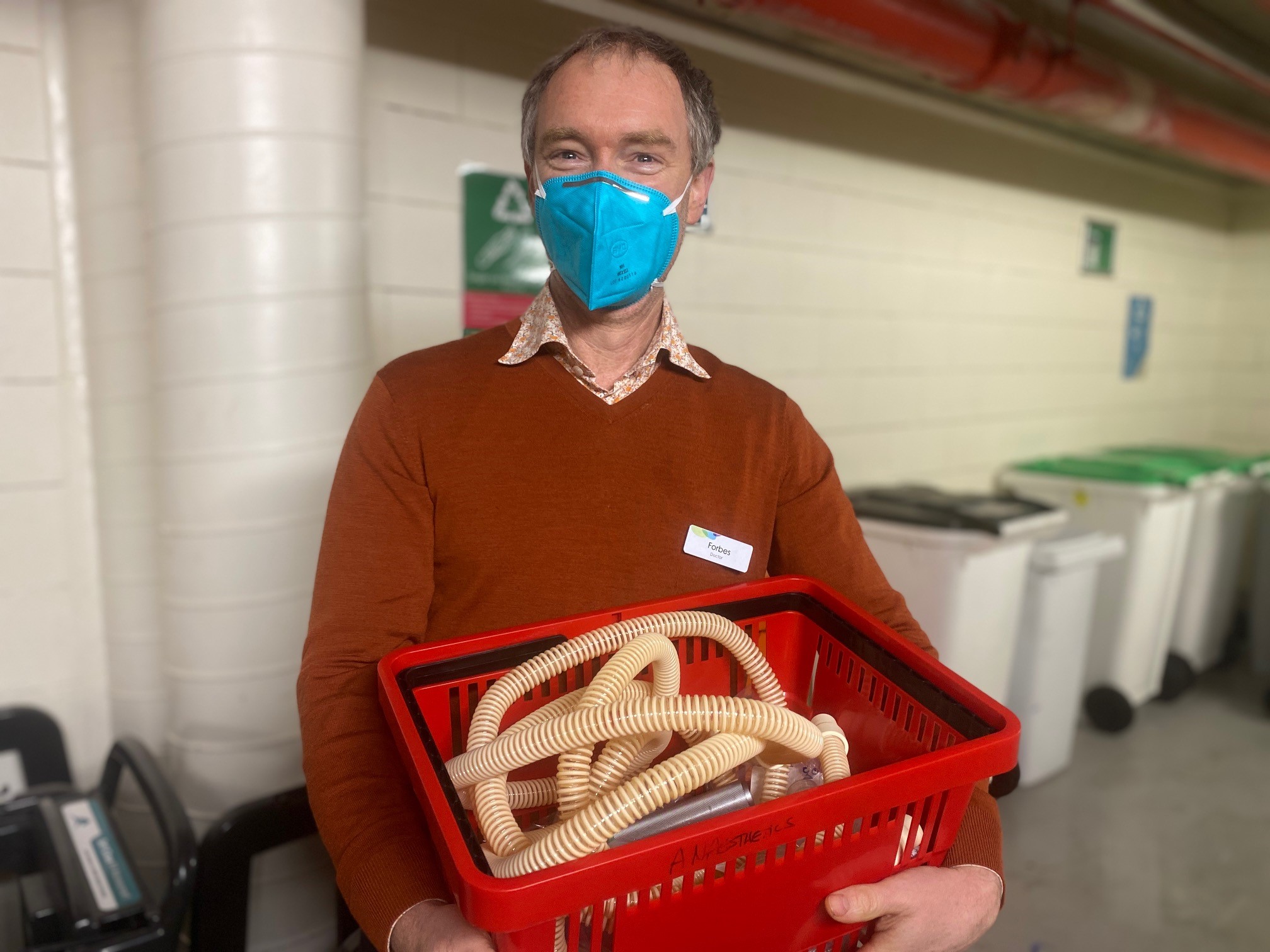 A man in a brown sweater holds a red bucket with a mask on.