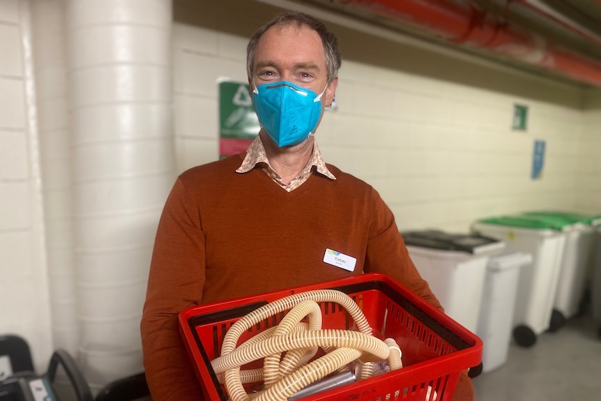 A man in a brown sweater holds a red bucket with a mask on.