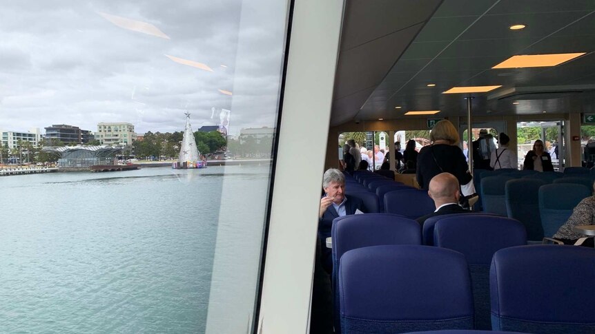 Passengers sit inside a modern ferry. The Geelong foreshore can be seen out the big windows.