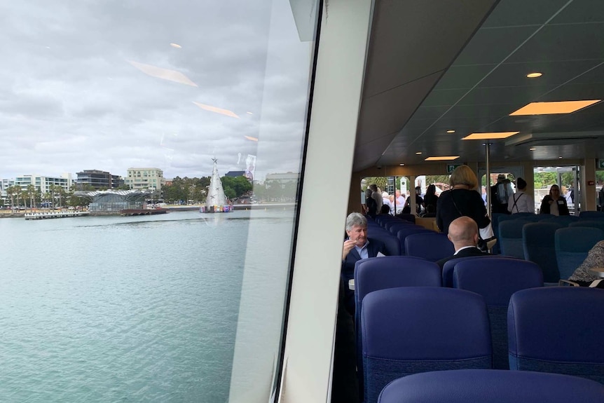 Passengers sit inside a modern ferry. The Geelong foreshore can be seen out the big windows.