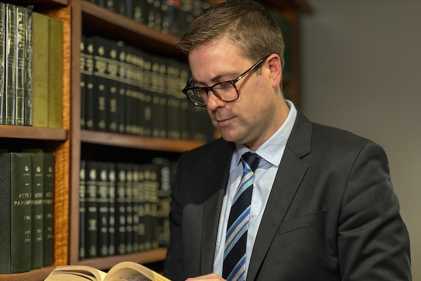 A man wearing a suit and glasses reads a book in front of a large bookshelf