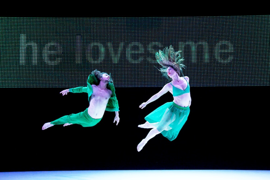 Two dancers wearing green leap into the air on a stage