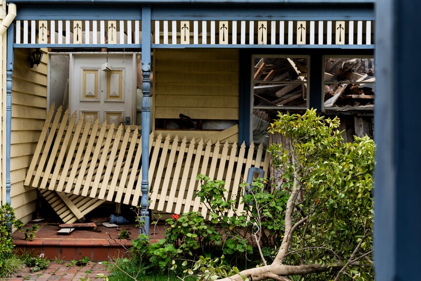 The front porch of a house undergoing demolition, rubble can be seen through the window and a broken fence sits by the door.