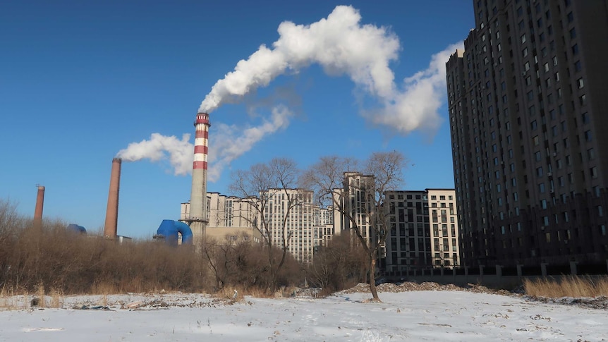 On a blue day, you view a snow-covered empty plot of land between high-rises with a chimney in the distance spewing white smoke.