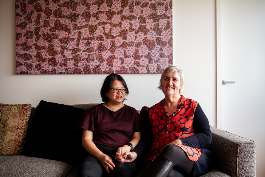 You view two women sitting on a grey patterened couch in front of an Australian Indigenous dot painting pictured on the wall.