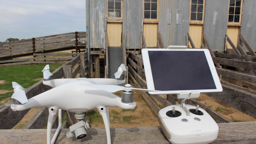 A white drone and control panel sits on a wooden rail in front of a shearing shed.