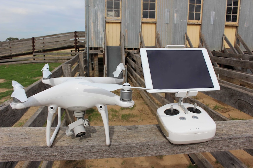 A white drone and control panel sits on a wooden rail in front of a shearing shed.