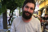 Axel Boreski sits at a table outdoors at night smiling with a plate of food in front of him holding a knife and fork.