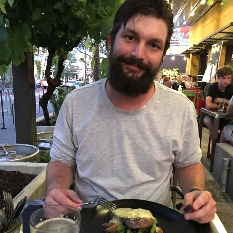 Axel Boreski sits at a table outdoors at night smiling with a plate of food in front of him holding a knife and fork.