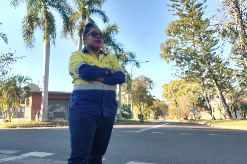 Sophie in a work high vis shirt, arms crossed, glasses on, palm trees and blue sky in the background.