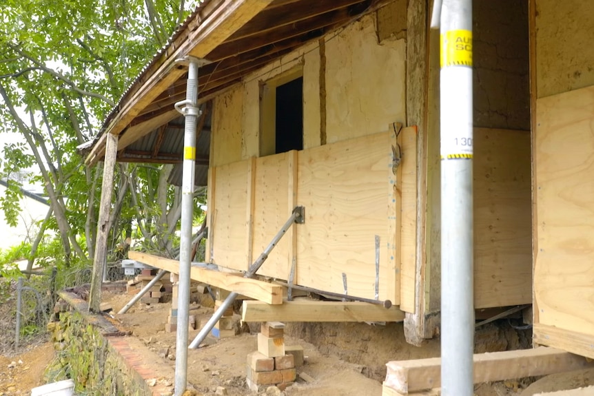 The front of a cottage which is being held up on timber beams underneath the floor. The walls are braced with plywood