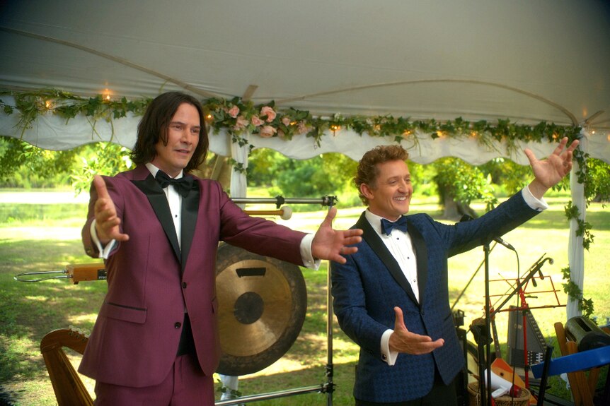 In outdoor tent on sunny day a dark haired man in maroon tux and shorter man in navy houndstooth suit stand with open arms.