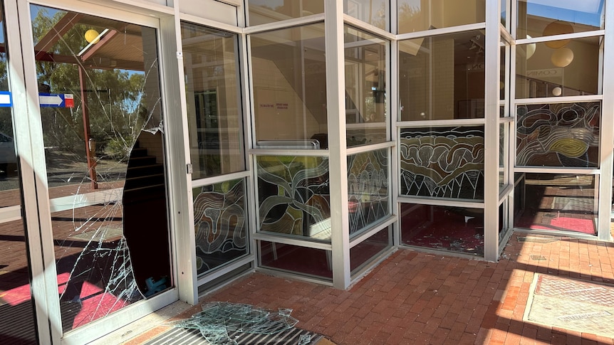 A glass door with glass windows surrounding is smashed. Shattered glass is on the ground. 