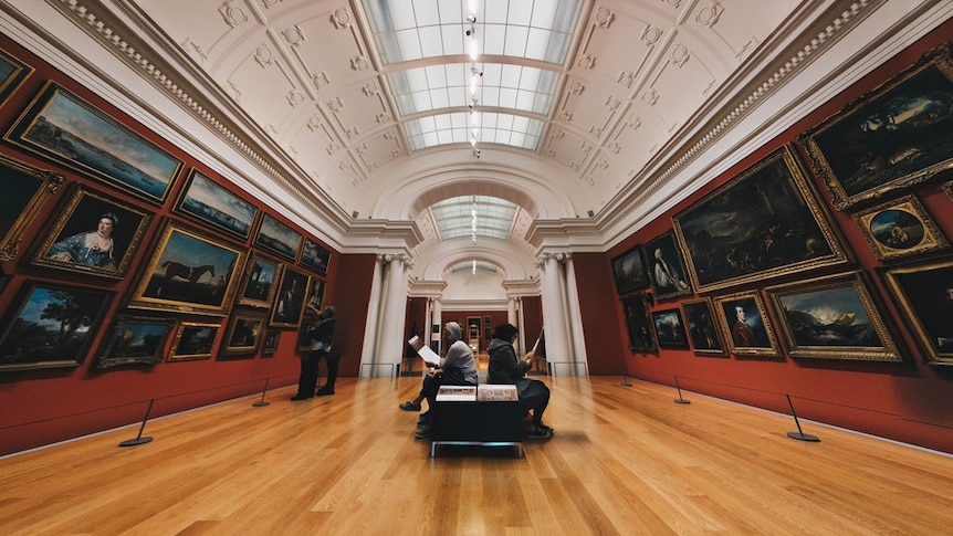 Two people sitting looking at catelogues in the middle of a large gallery with walls covered in paintings.