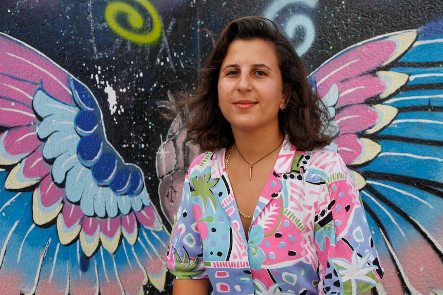 A woman in a colourful shirt sits in front of a mural with a slight smile on her face