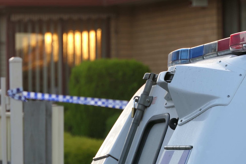 A police car in focus and a home with police tape in front of it in the morning light in the background.