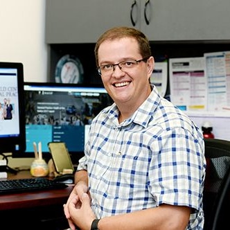 A man with glasses and a checked shirt sitting in front of a computer screen.