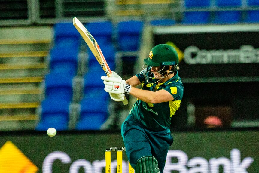 A medium shot of a female cricketer with a helmet on holding up their bat while striking a ball during a match.