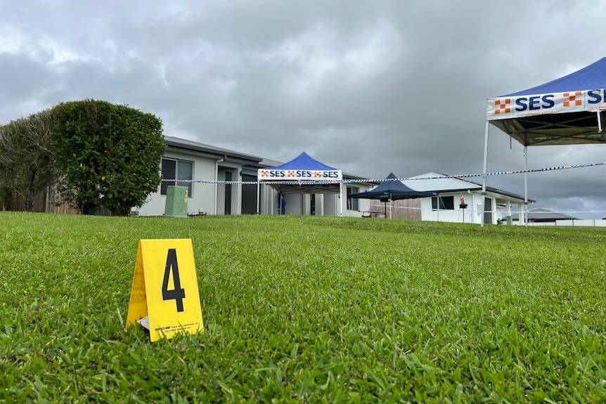 A little yellow marker with the number 4, likely to mark evidence, on pristen green lawn. In the background police tents.