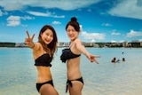 Two women in swimsuits smile at the camera on the beach