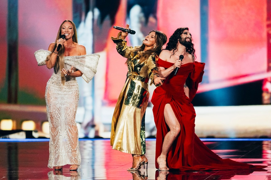 Three people on stage wearing dresses, performing, one has a beard and long hair
