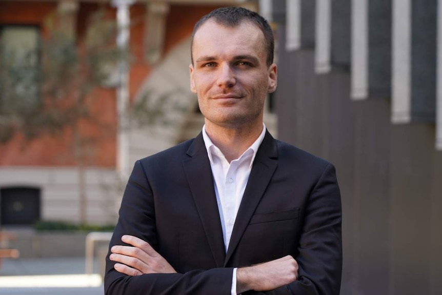 A man in a suit stands with his hands crossed in a city location.