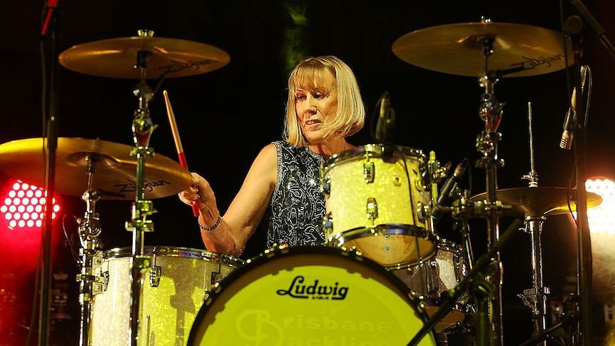 Lindy Morrison, with shoulder-length blonde hair, in dark room and with neutral expression, playing the drums.