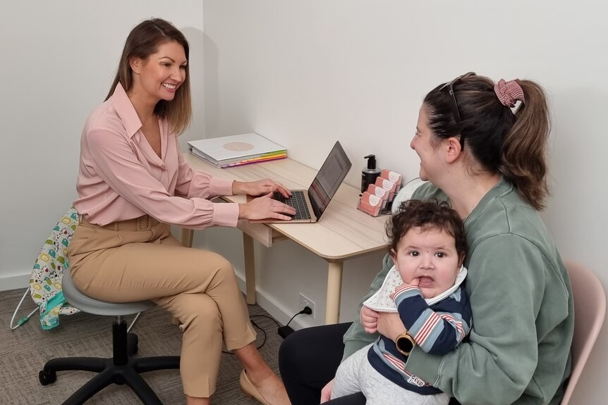 Two women sit together in an office, one holds a baby.