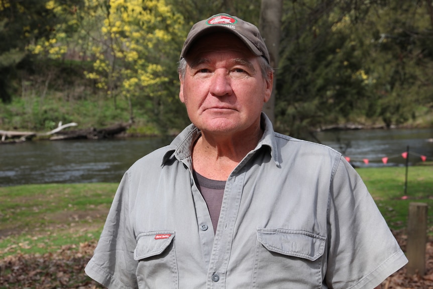 An elderly man, wearing a Rabbitohs cap, standing in front of a flowing river.