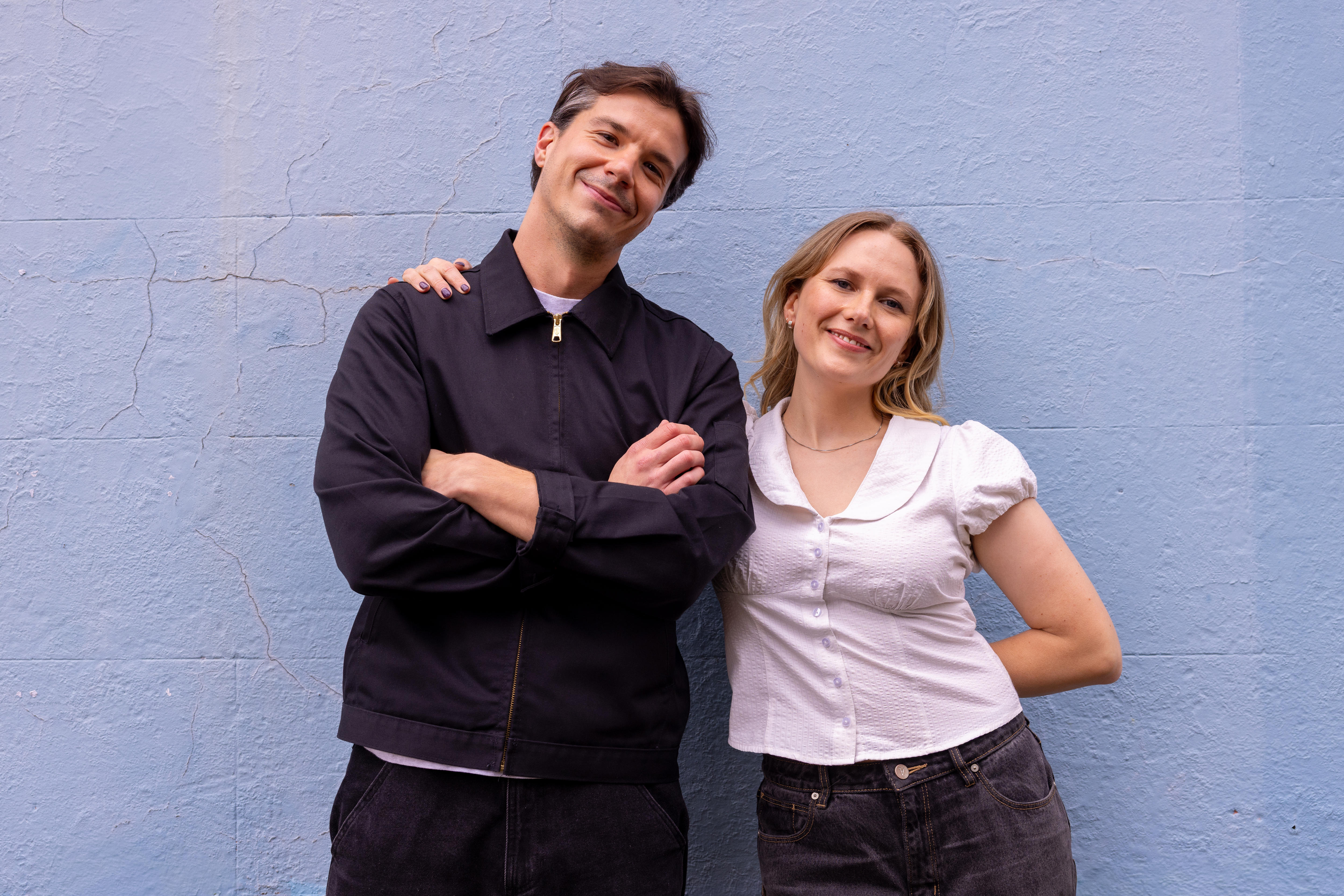A man in a dark zip up jacket stands next to a shorter blonde woman in a white top against a grey wall