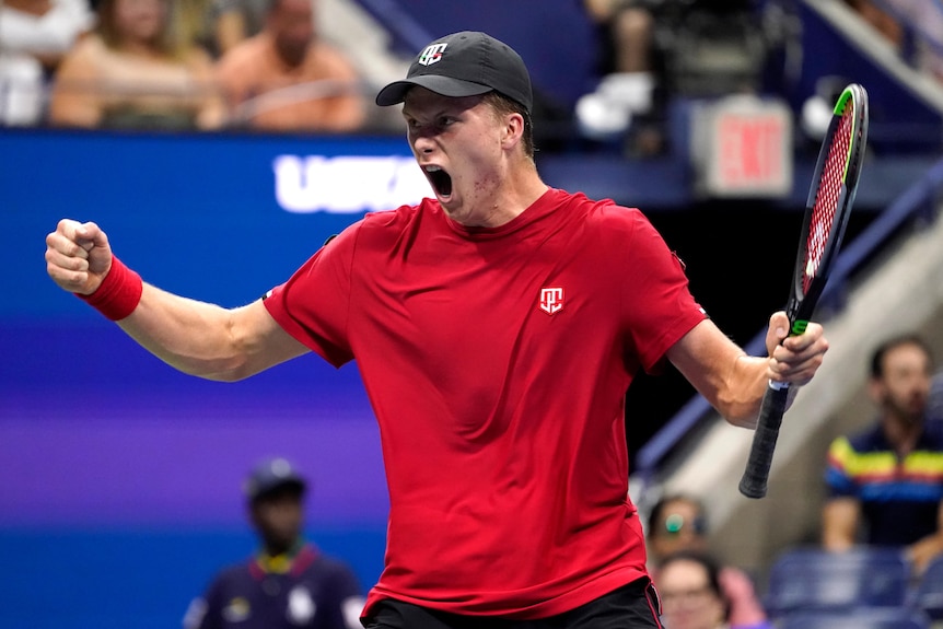 Jenson Brooksby, in a black cap and red shirt, holds a tennis racquet as he stretches out his arms and shouts at the US Open.