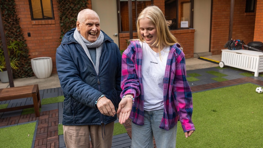 old bald man smiling with his hand extended alongside a blonde haired girl smiling with her hand out