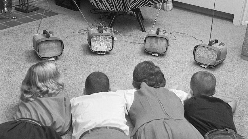 Black and white photo of four children on the floor watching four small television sets.