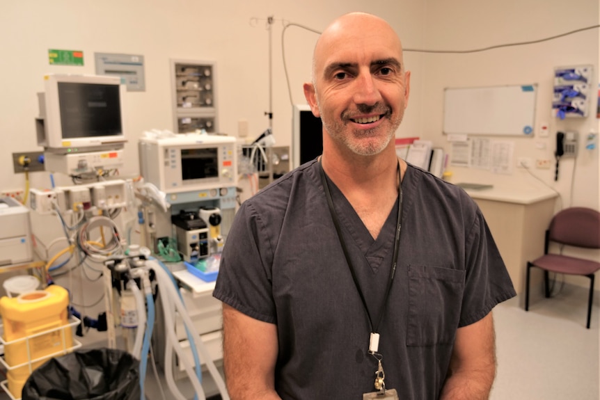 Man standing in the middle of an operating theatre with surgical machinery behind him.